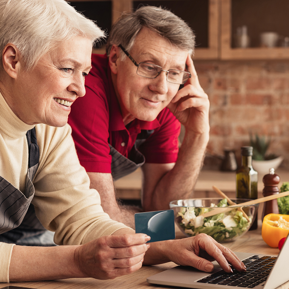 Ordering Food Online. Smiling elderly couple with laptop and credit card purchasing grocery delivery from internet while cooking together in kitchen